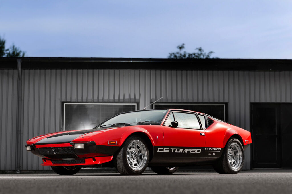 De Tomaso Pantera GTS i röd och svart lack, fotograferad i profil framför industriell byggnad.