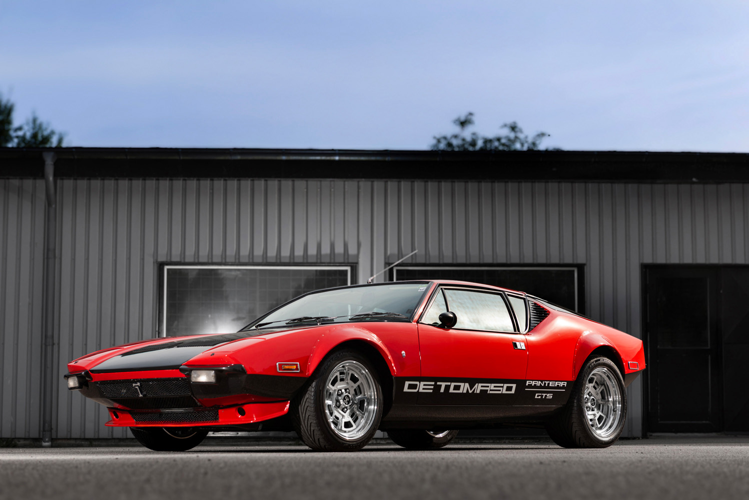 De Tomaso Pantera GTS i röd och svart lack, fotograferad i profil framför industriell byggnad.